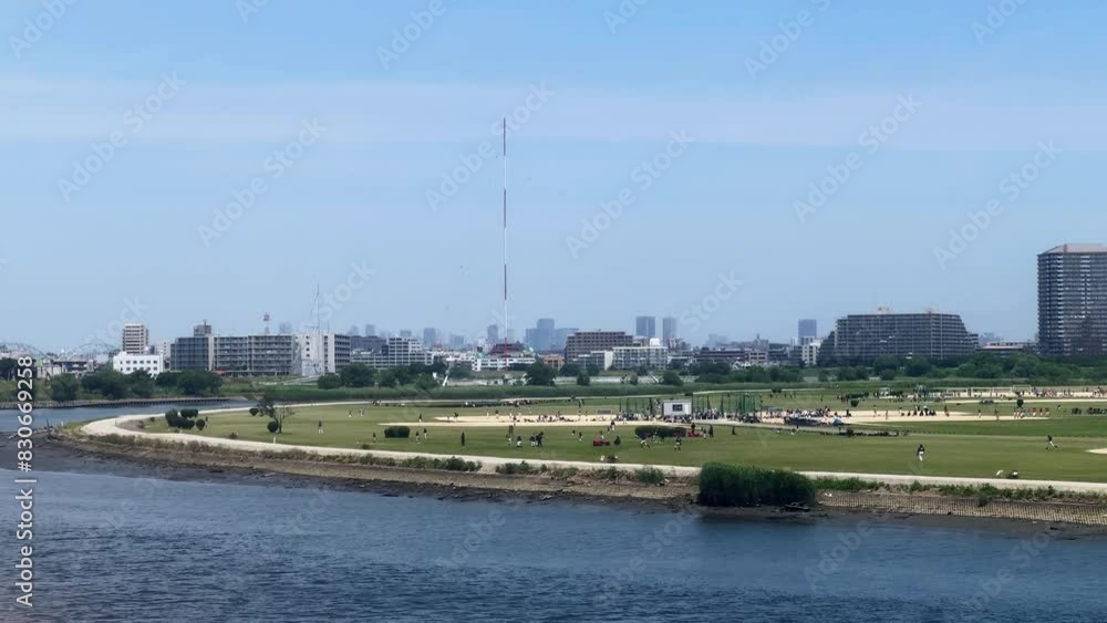 Park near river on sunny day with cityscape in the background, people enjoying outdoor activities Park near river on sunny day with cityscape in the background, people enjoying outdoor activities