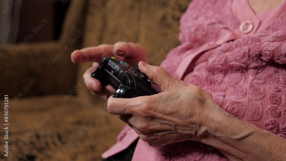 Close-up of an elderly grandmother with wrinkled hands holding a joystick, she is playing video games at home sitting on the couch. Close-up of an elderly grandmother with wrinkled hands holding a joystick, she is playing video games at home sitting on the couch.