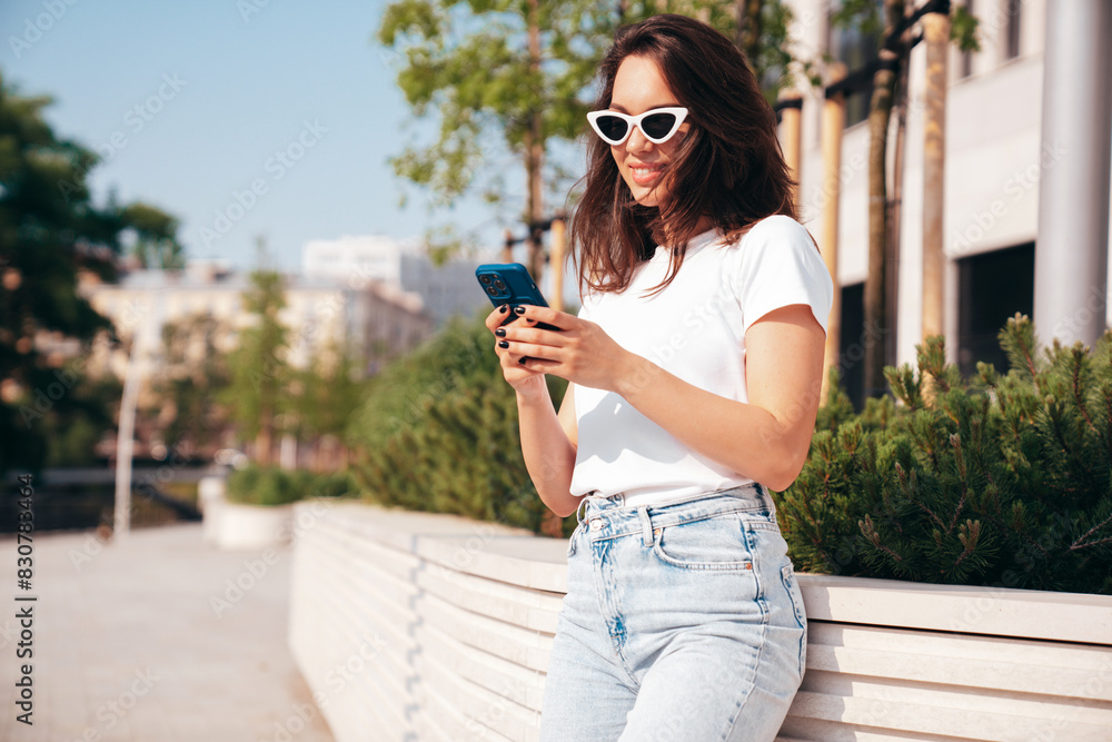 Young beautiful smiling hipster woman in trendy summer clothes. Carefree model posing in street at sunset. Positive female. Holds smartphone, uses phone apps, looks at telephone screen Young beautiful smiling hipster woman in trendy summer clothes. Carefree model posing in street at sunset. Positive female. Holds smartphone, uses phone apps, looks at telephone screen