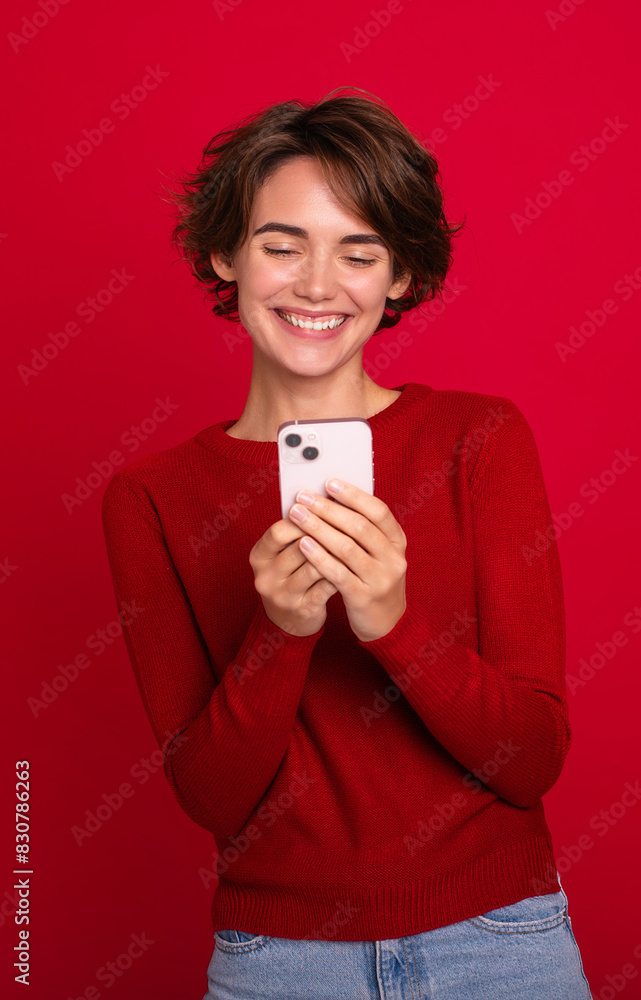 Naklejka premium Vertical photo of a smiling beautiful young modern woman who using a smartphone isolated on a red bright wall. Shopping, chatting, surfing the internet, betting, social media, win in games
