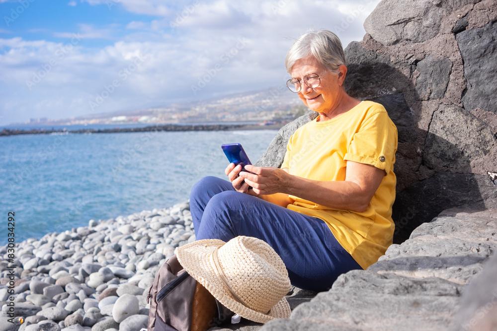 Attractive senior woman sitting on a pebble beach at sea using mobile phone. Mature woman smiles holding cellphone in hand. Concept of vacation, freedom and happy retirement