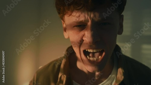 Pull in portrait of stressed out Caucasian man screaming through pain and looking at camera in dimply lit room at night photo