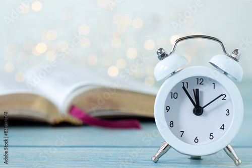 Alarm clock and open holy bible book with white bokeh light background. Close-up. Copy space. Christian biblical concept of patience, time, prayer, and Scripture study.