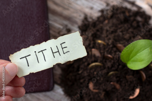 Hand holding handwritten word "tithe" on top of soil with golden coin money and green plant. Close-up. Christian biblical concept of tithing, generosity, obedience to God Jesus Christ.