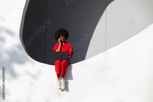 Mature woman in red jumpsuit with laptop talking through smart phone sitting on white wall