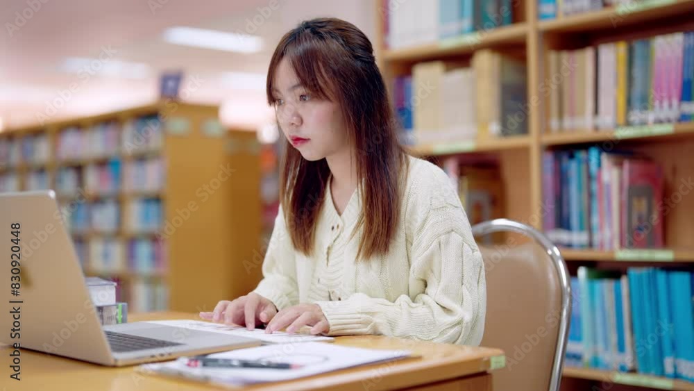 A woman is sitting at a desk in a library, typing on a laptop. She is focused and serious, possibly working on a project or studying. The library setting suggests a quiet and studious atmosphere A woman is sitting at a desk in a library, typing on a laptop. She is focused and serious, possibly working on a project or studying. The library setting suggests a quiet and studious atmosphere