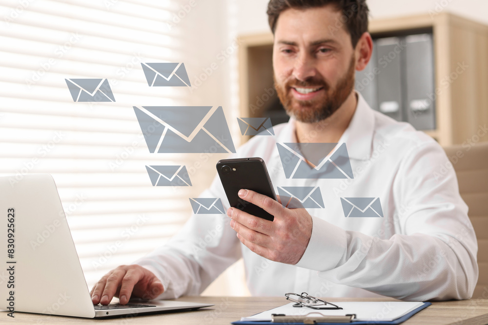 Smiling man with smartphone chatting at table in office. Many illustrations of envelope as incoming messages around device Smiling man with smartphone chatting at table in office. Many illustrations of envelope as incoming messages around device