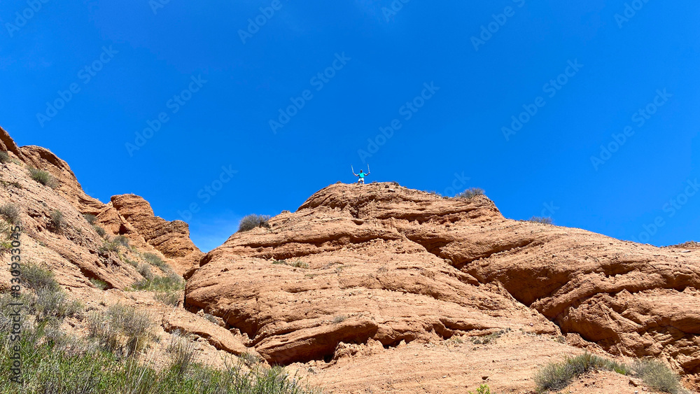 Fototapeta premium A man stands on top of a rocky hill, admiring the scenery. The red canyons of Konorchek. Travel to Kyrgyzstan. The sky is clear and blue