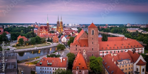 Stunning aerial view of Wrocław's historic Cathedral Island (Ostrów Tumski) at dusk, showcasing the beautifully illuminated gothic architecture, red-tiled roofs and serene river.