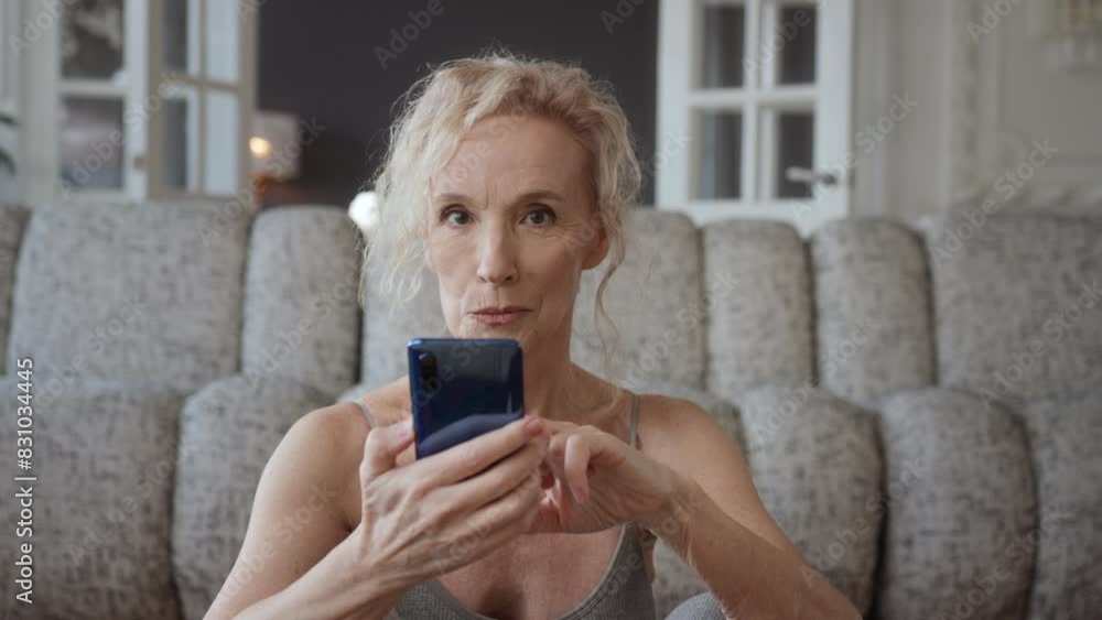 Woman in 60s sitting on chair at home with legs on windowsill, relaxing and using mobile phone. Enjoys leisure browsing social media or online shopping. Satisfaction and fulfillment in woman life. Woman in 60s sitting on chair at home with legs on windowsill, relaxing and using mobile phone. Enjoys leisure browsing social media or online shopping. Satisfaction and fulfillment in woman life.