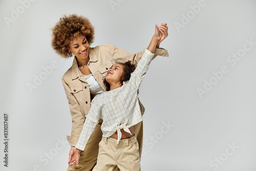 A curly African American mother and daughter joyfully dancing in stylish outfits on a gray background.