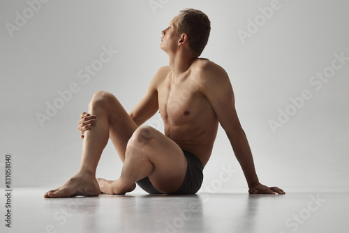 Young bode guy sitting in underwear shirtless with thoughtful expression isolated on grey studio background. Defines physique. Concept of male beauty, body care, fitness, sport, health photo