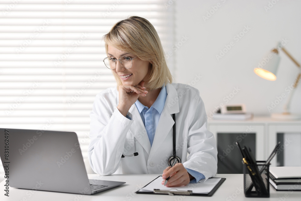 Smiling doctor with laptop having online consultation at table in office Smiling doctor with laptop having online consultation at table in office
