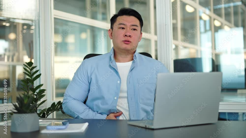 Frustrated chinese male expressing stomach ache while working in corporate office environment. Upset businessman dressed in casual blue shirt massaging belly indoors. Concept of suffering and ill. Frustrated chinese male expressing stomach ache while working in corporate office environment. Upset businessman dressed in casual blue shirt massaging belly indoors. Concept of suffering and ill.