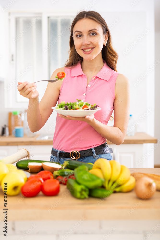 Cheerful svelte young woman eating vegetable salad in home kitchen Cheerful svelte young woman eating vegetable salad in home kitchen