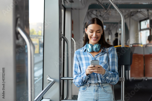 Happy European woman with headphones using cellphone, training in modern tram, enjoying comfortable public transport. Portrait of passenger with phone and headset