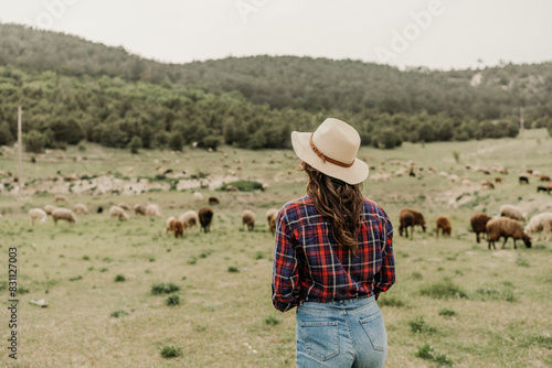 A woman wearing a straw hat stands in a field with a herd of sheep