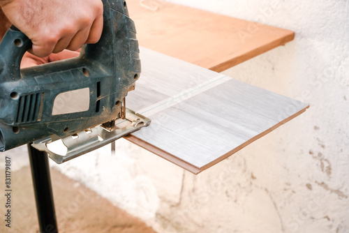 Close up of male construction worker hands cutting laminate wooden board with jigsaw tool. Man using electric power saw cutter while preparing laminate material for floor installation