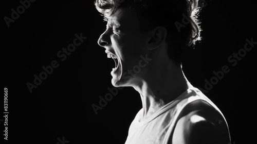 Monochrome studio portrait in profile of furious young man in undershirt shouting at top of his voice against black background photo