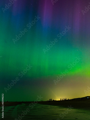 People at the beach are watching a majestic display of colorful Northern Lights over the North Sea.