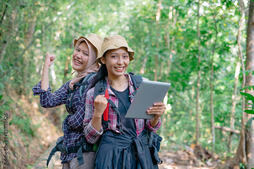 Two beautiful Asian backpacker female holding tablet and rising clenched fist at the forest 