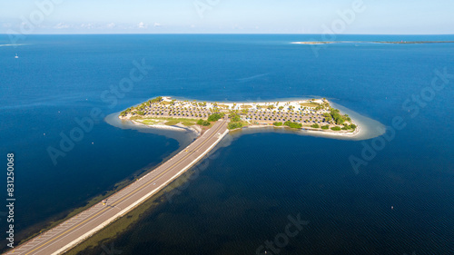 Aerial view of Fred Howard Park in Tarpon Springs, Florida, featuring parking lots, palm trees, sandy beaches, and surrounding blue waters under a clear sky. photo