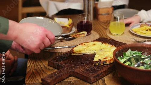 Unrecognizable person taking piece of khachapuri with meat and cheese using a metal multifunctional device. Traditional georgian dish served on thick wooden board on table with plates and drinks.