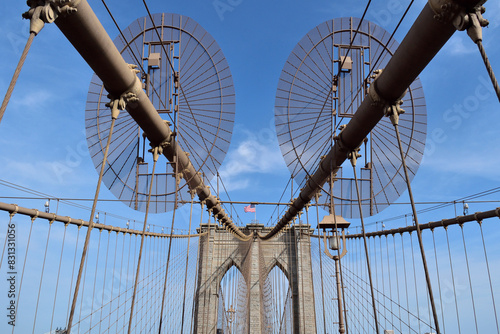 Brooklyn bridge is a large, metal structure with two large, circular wheels on top. The sky is clear and blue, and there is a flag flying above the bridge