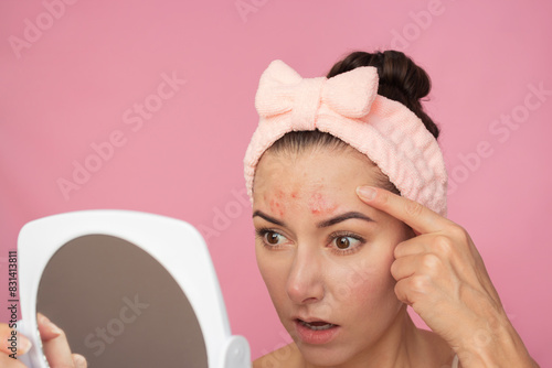 Young pretty caucasian brunette woman with a pink headband looks at a white mirror, she is upset, saw pimples on her forehead . Isolated on a pink background photo