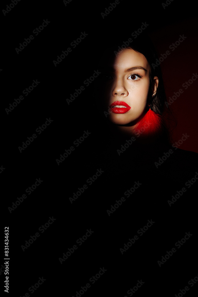 The face of a model girl in a spot of light on a black photo. Portrait of a model with bright makeup and red lipstick. The face of a model girl in a spot of light on a black photo. Portrait of a model with bright makeup and red lipstick.