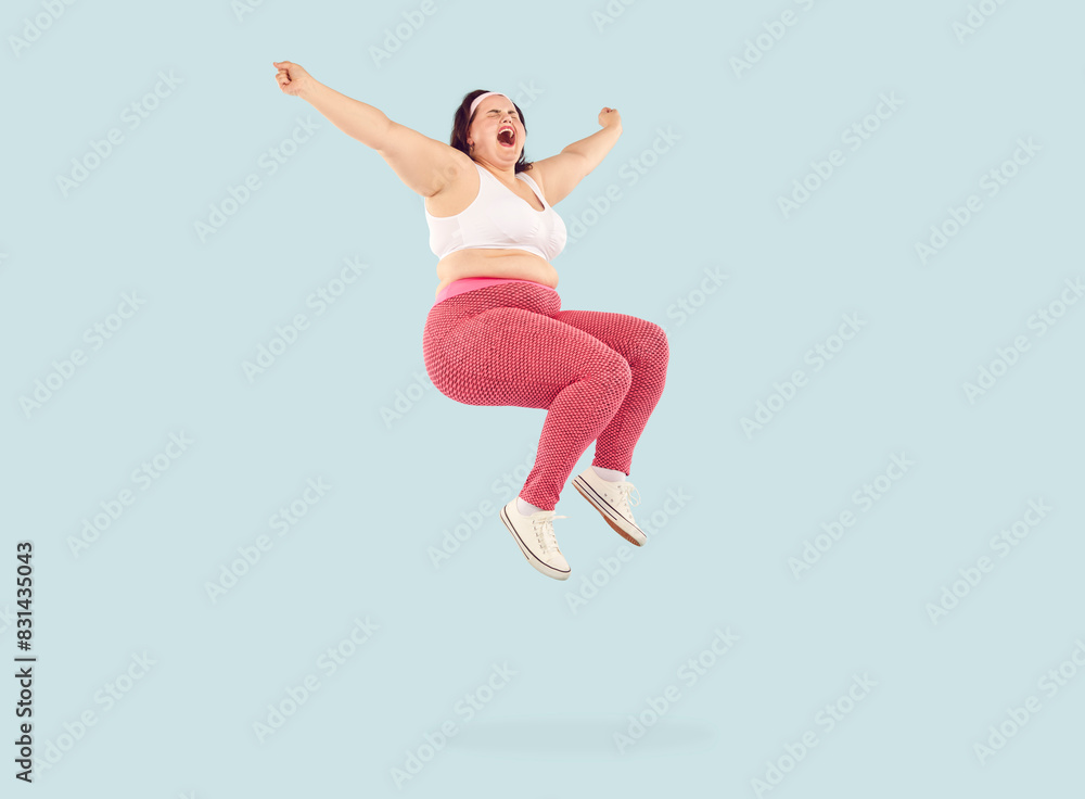 Fototapeta premium Full length shot of funny chubby overweight woman jumping in air. Happy plus size, fat woman wearing sportswear jumping high with her hands outstretched isolated on light blue studio background