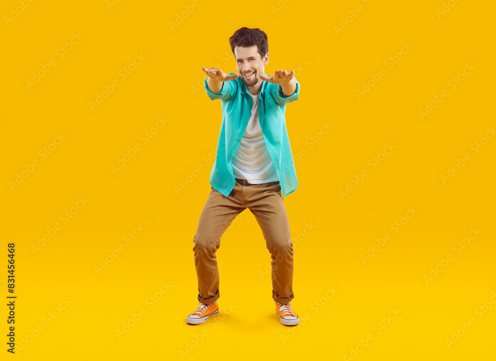Cheerful funny guy jokingly pretending to squat with arms outstretched, isolated on orange background. Young Caucasian man in casual shirt, t-shirt and pants is fooling around and smiling. Full length Cheerful funny guy jokingly pretending to squat with arms outstretched, isolated on orange background. Young Caucasian man in casual shirt, t-shirt and pants is fooling around and smiling. Full length