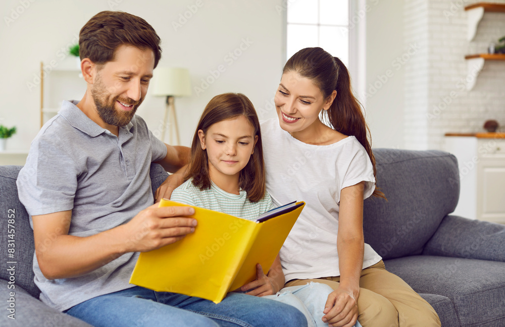 Young happy family reading a book with their little cute daughter sitting on sofa at home. Smiling joyful parents looking through a family photo album with their child girl. Family leisure concept. Young happy family reading a book with their little cute daughter sitting on sofa at home. Smiling joyful parents looking through a family photo album with their child girl. Family leisure concept.