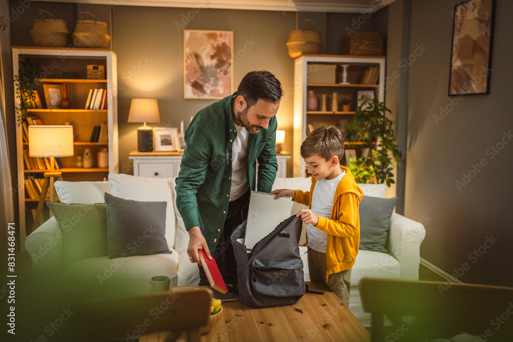 Dad help his son to pack his notebook and book for school in backpack Dad help his son to pack his notebook and book for school in backpack