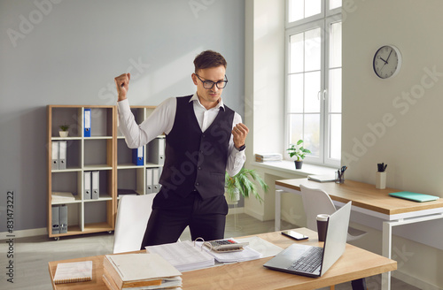 Fotografie Happy emotional confident accountant standing by office desk with computer raising fists up