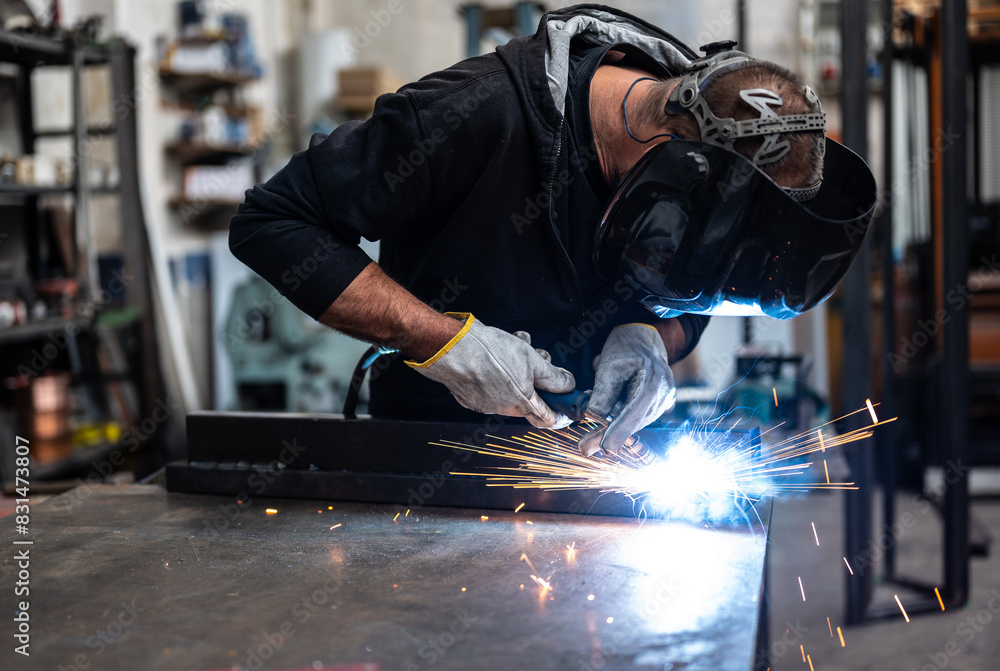 Performing a weld: the man in the welding mask is positioned with the welding machine on the welding point. When he activates it, a blue glow with smoke and orange sparks illuminates the scene. Performing a weld: the man in the welding mask is positioned with the welding machine on the welding point. When he activates it, a blue glow with smoke and orange sparks illuminates the scene.