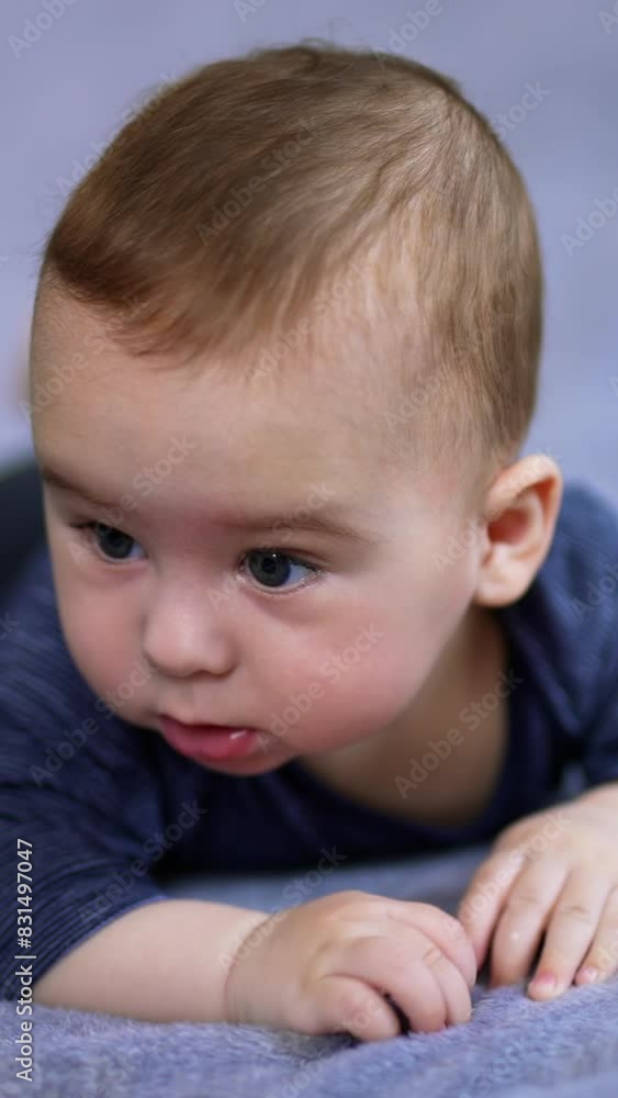 Beautiful child lies on belly moving his feet playfully. Adorable baby boy in blue clothes takes his hand into mouth. Close up. Blurred backdrop. Vertical video Beautiful child lies on belly moving his feet playfully. Adorable baby boy in blue clothes takes his hand into mouth. Close up. Blurred backdrop. Vertical video