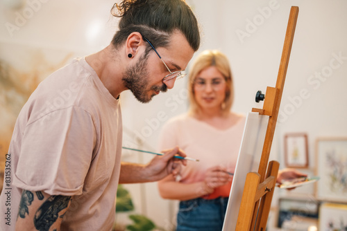 Creative artist painting on easel at art studio with woman in background.