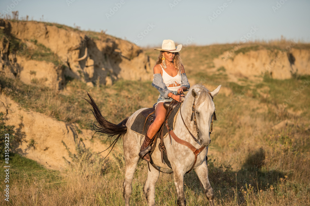 Sexy cowgirl with hat on horseback in the desert. Beautiful girl in a cowboy hat and denim on a horse. Beautiful cowgirl on horseback in the mountains. Outdoor lifestyle portrait