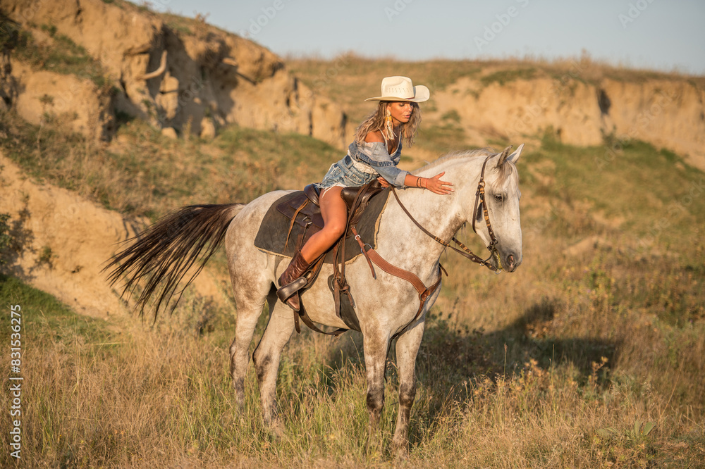 Sexy cowgirl with hat on horseback in the desert. Beautiful girl in a cowboy hat and denim on a horse. Beautiful cowgirl on horseback in the mountains. Outdoor lifestyle portrait