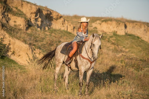 Obraz na płótnie Sexy cowgirl with hat on horseback in the desert. Beautiful girl in a cowboy hat and denim on a horse. Beautiful cowgirl on horseback in the mountains. Outdoor lifestyle portrait w salonie