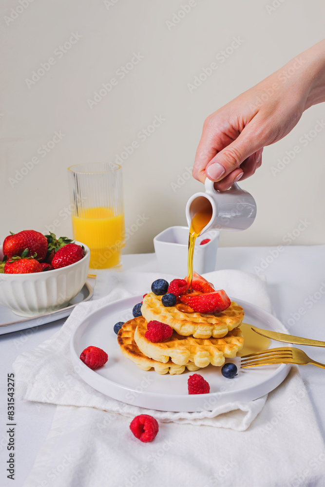 Close-up of the hand pouring maple syrup from the saucer on top of sweet homemade waffles with berries Close-up of the hand pouring maple syrup from the saucer on top of sweet homemade waffles with berries