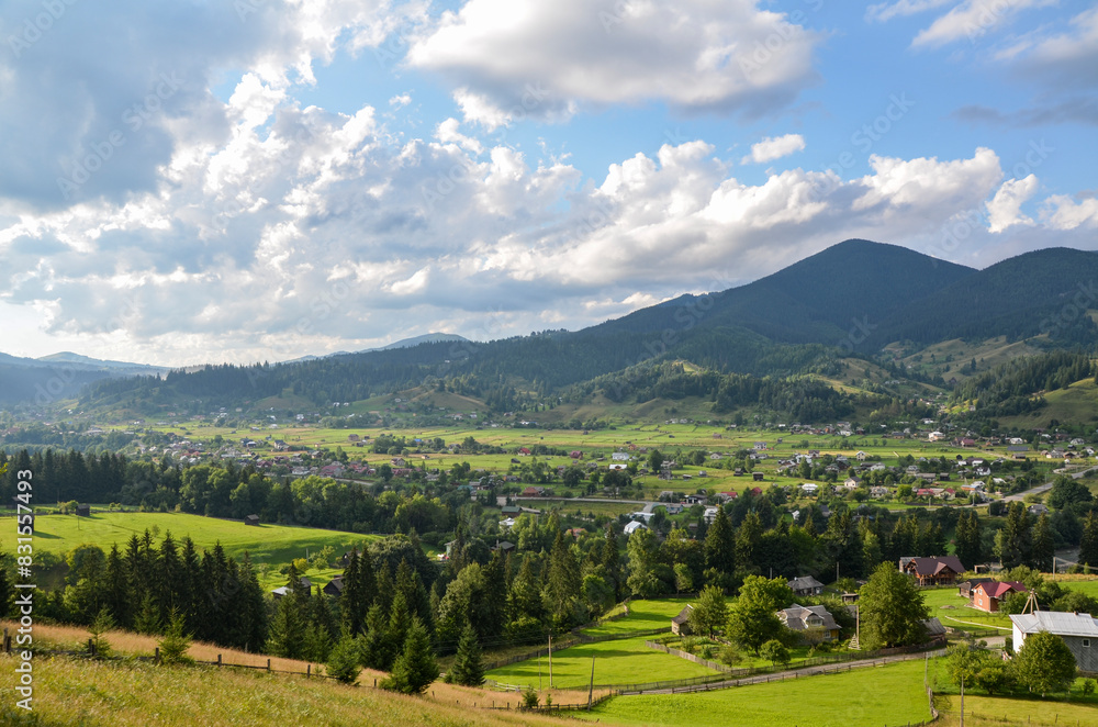 Naklejka premium Rural landscape is adorned with scattered houses, verdant trees, and majestic mountains with patches of forests in the background. Verkhovyna, Carpathian Mountains, Ukraine