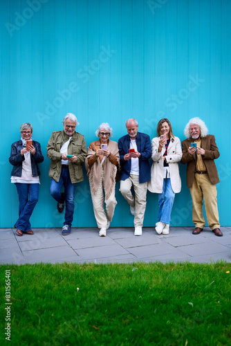 Vertical. Group of elderly Caucasian smiling together using cell phone leaning on blue wall. Happy old friends gathered having fun looking mobile outdoor. Mature people enjoying devices. Copy space