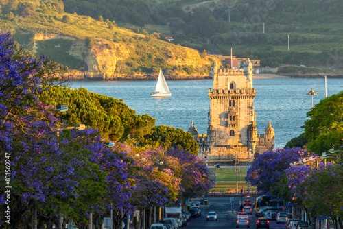Belem Tower, Jacaranda Blooming Purple Blue Trees and Sailboat on Sunny Evening. Golden Hour. Lisbon, Portugal.