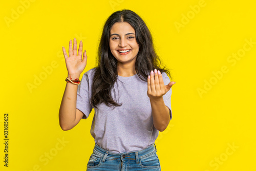 Young Indian woman smiling friendly at camera, waving hands gesturing hello greeting or goodbye welcoming with invitation hospitable expression. Arabian Hindu girl isolated on yellow studio background