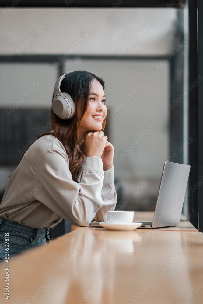 Obraz premium A woman wearing headphones is sitting at a table with a laptop and a cup of coffee. She is smiling and she is enjoying her time