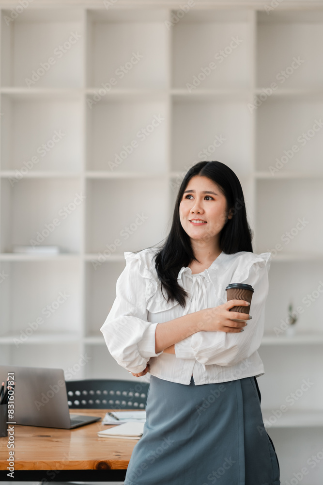 A woman in a white shirt and blue skirt is holding a coffee cup. She is standing in front of a white shelf with a laptop on it A woman in a white shirt and blue skirt is holding a coffee cup. She is standing in front of a white shelf with a laptop on it