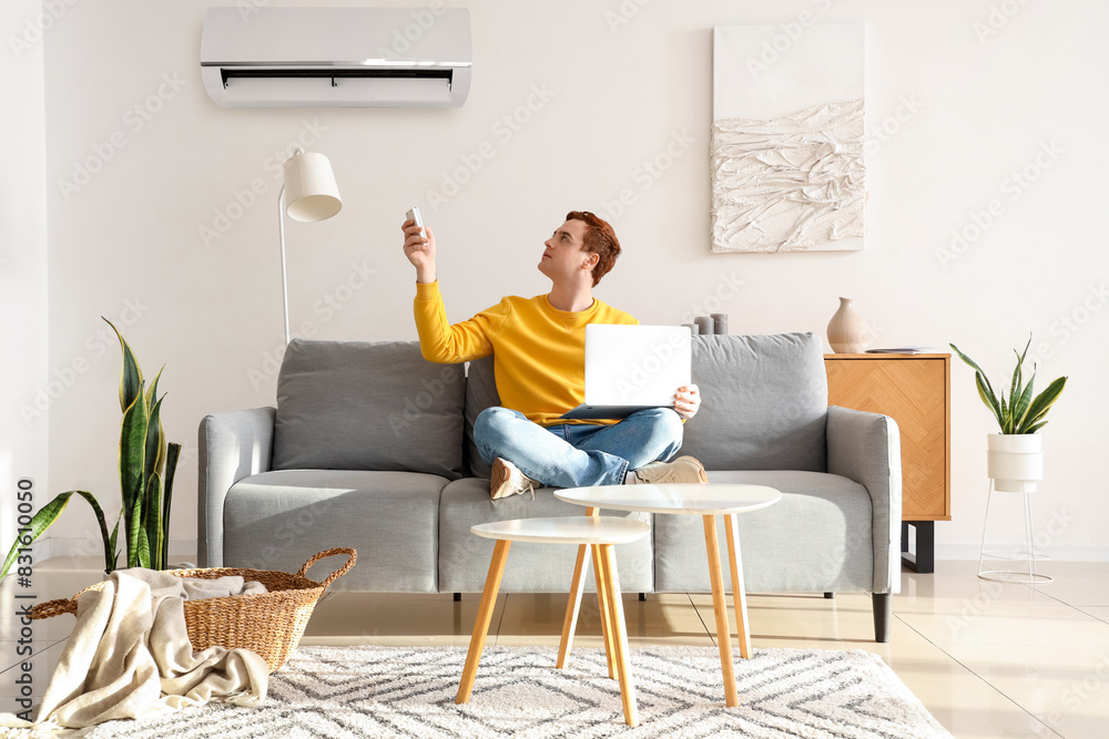 Young man with laptop turning on air conditioner on couch in living room Young man with laptop turning on air conditioner on couch in living room