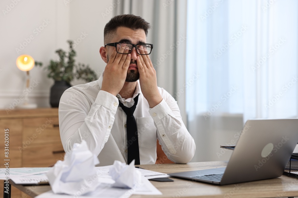 Overwhelmed man sitting at table with laptop and documents in office Overwhelmed man sitting at table with laptop and documents in office